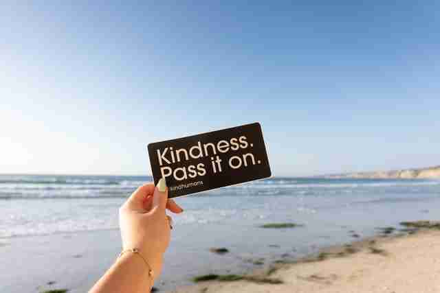 woman's hand holding a sign that says Kindness. pass it on. while standing on a beach with the ocean in a backround