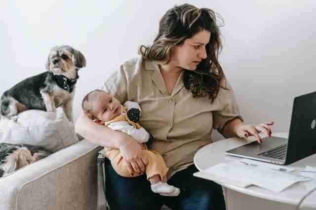 woman being very effective by multitasking with a baby in one arm, typing on a laptop with another while also watching her pet dogs